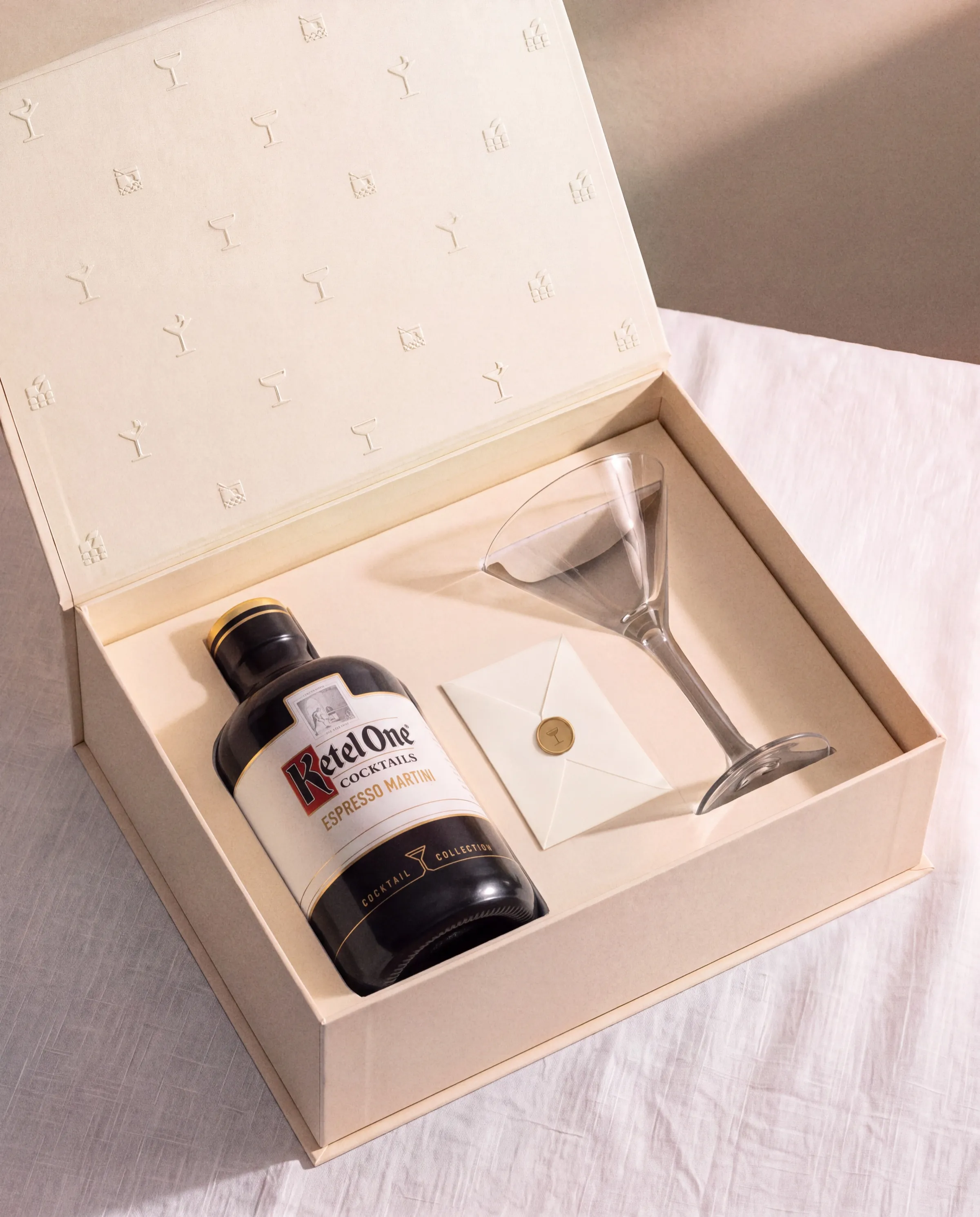 The Cocktail Collection gift box, open: an embossed cream lid, a Ketel One Espresso Martini bottle, a sealed envelope, and a martini glass