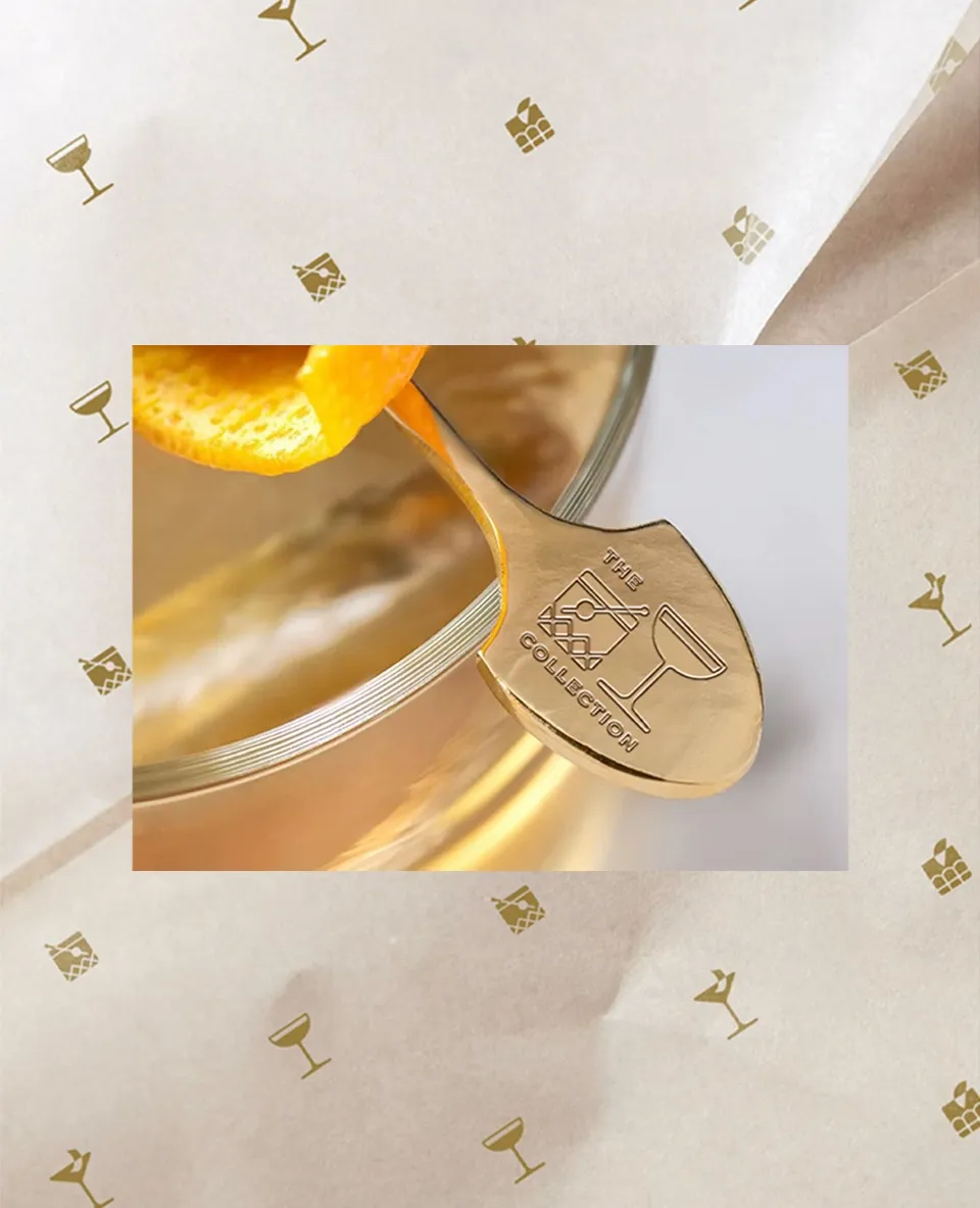 A gold Cocktail Collection cocktail pick with an orange twist, on the icon-patterned tissue paper