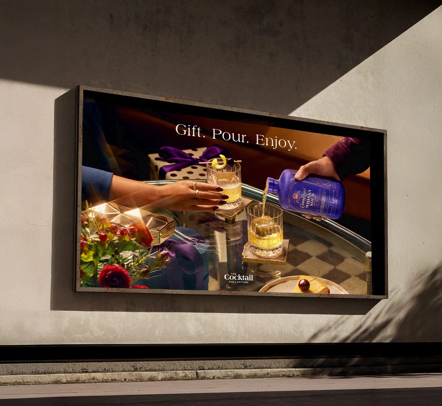 The "Gift. Pour. Enjoy." holiday billboard at street level: Crown Royal Whisky Sour poured over ice beside a wrapped gift, The Cocktail Collection