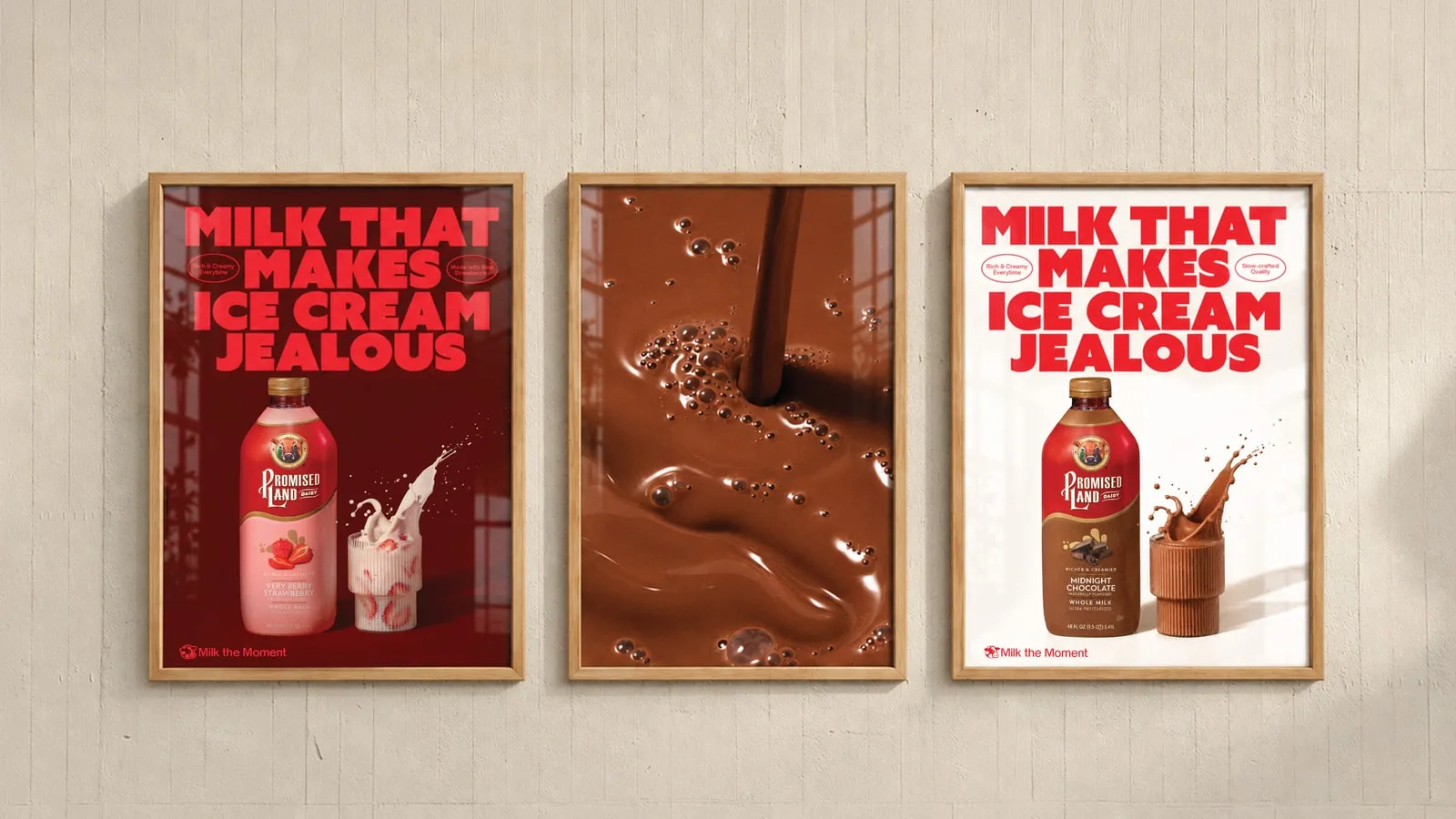 Three framed posters on a pale wooden wall: two read “Milk that makes ice cream jealous”, one with a Promised Land Dairy Very Berry Strawberry bottle and one with a Midnight Chocolate bottle, both signed “Milk the Moment”; the middle poster is a macro of pouring chocolate milk